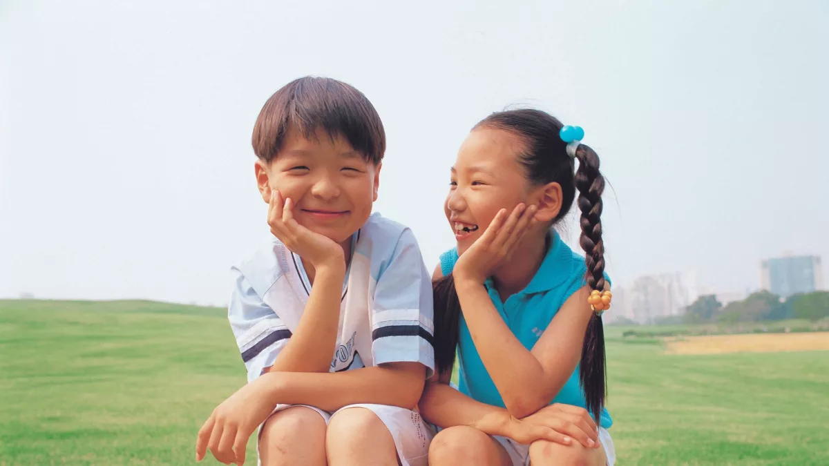 Duas crianças de etnia amarela sorriem sentados em um gramado. A imagem ilustra matéria sobre a atuação do Alana nas eleições de conselhos tutelares em 2023.