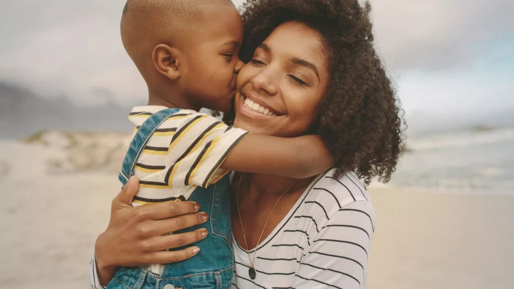 Uma família negra composta por mãe e filho. A mãe está sorridente e recebe um beijo da criança. A imagem ilustra a matéria "Instituto Alana aborda o direito ao cuidado sob a ótica das infâncias na Corte Interamericana de Direitos Humanos".