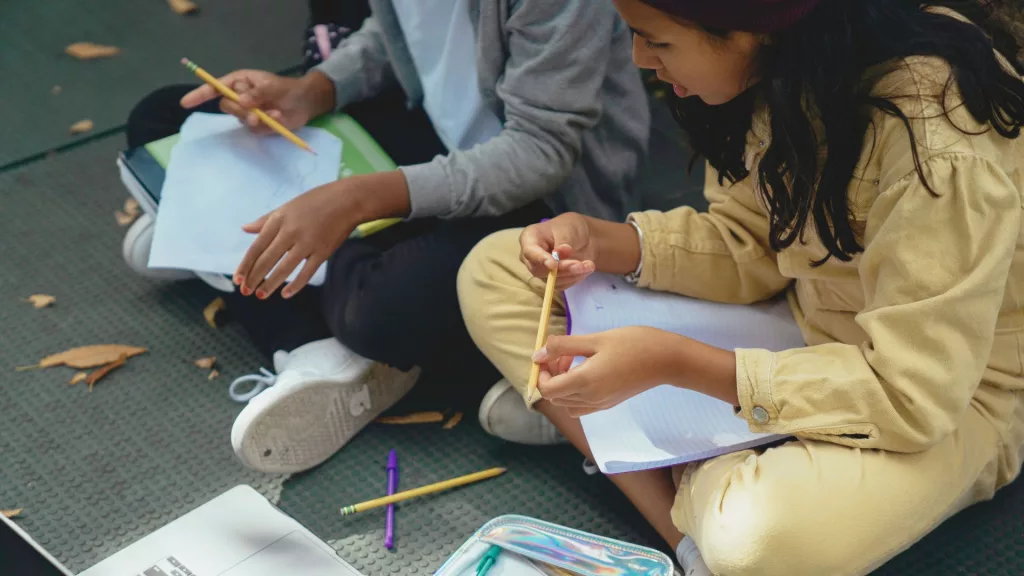 Na imagem, duas meninas estão sentadas no chão com cadernos e lápis nas mãos. A foto ilustra a matéria "Instituto Alana envia contribuição internacional destacando como a crise climática afeta os direitos de meninas"