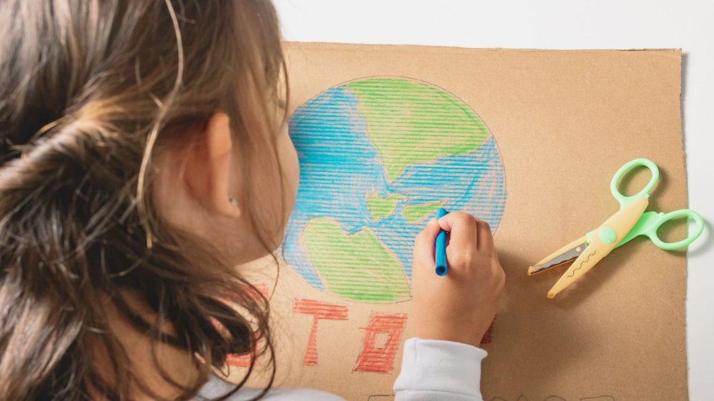 Uma menina desenha o planeta Terra em um papelão. A imagem ilustra a matéria "Audiência da Corte IDH aborda garantia de direitos e participação das infâncias em soluções para a emergência climática".
