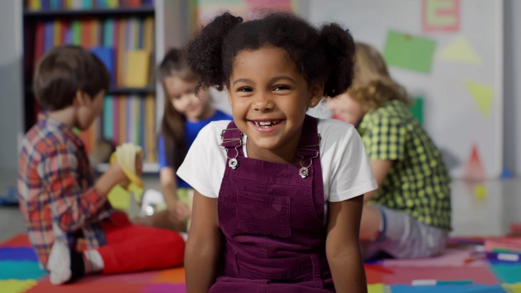 CNE: na imagem, uma menina negra sorri. Ela está em uma sala de aula. Ao fundo, outras crianças estão sentadas.