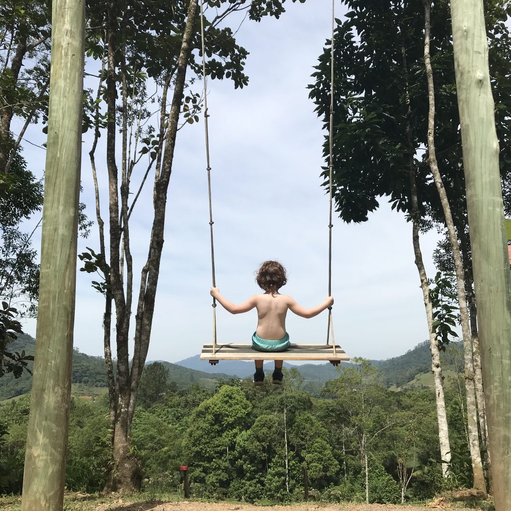 Em um balanço pendurado entre árvores, uma criança olha para um ambiente verde, de mata